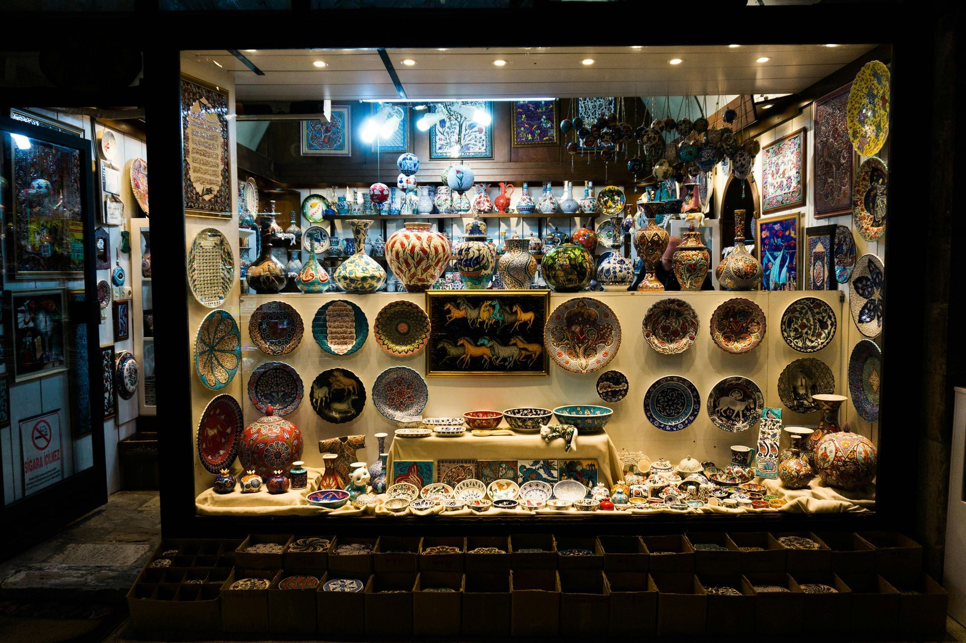 Shop window display filled with colorful ceramic plates, vases, and small trinkets, lit from above at night.