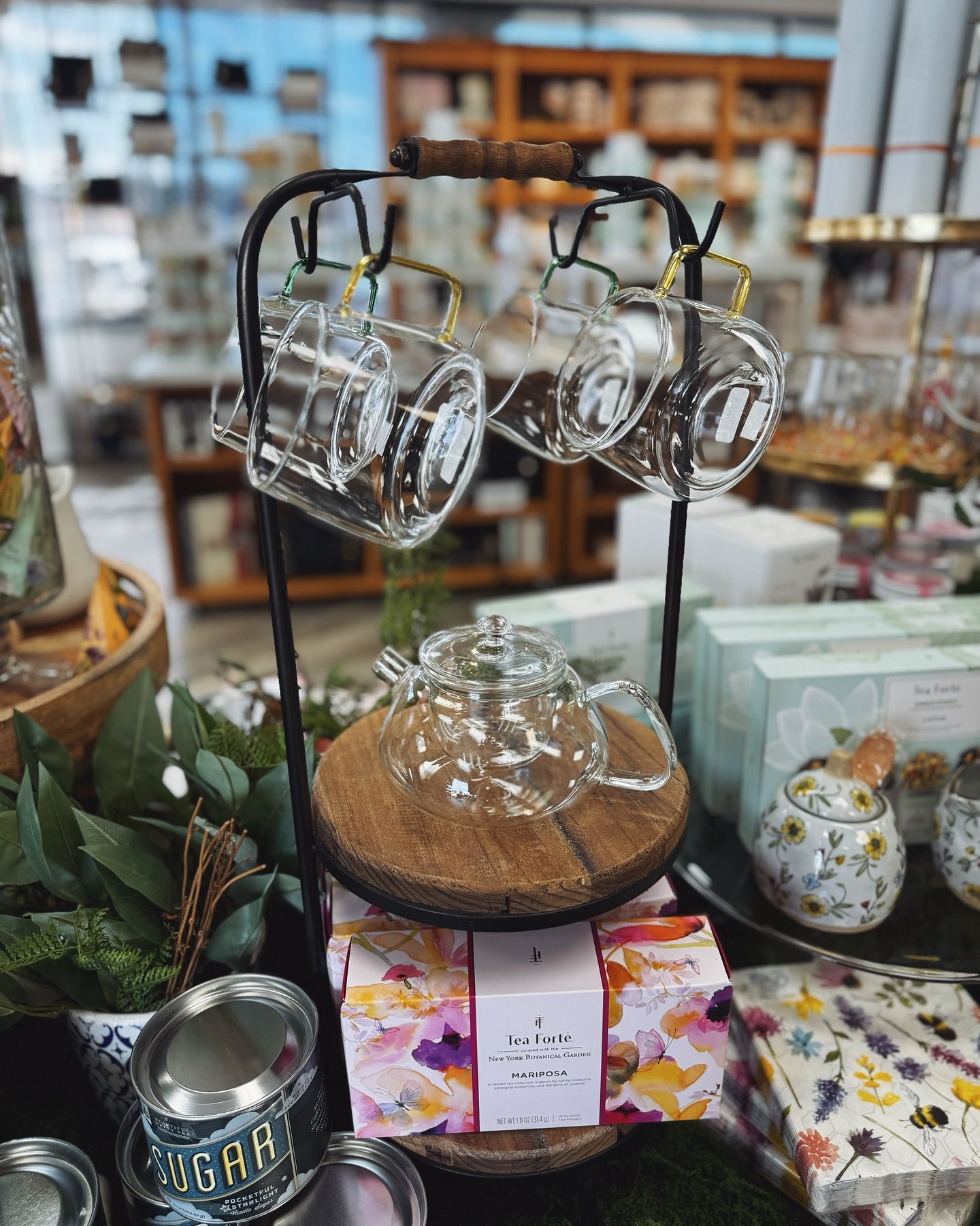 Glass teacups and teapot on a metal stand displayed in a store. Cork base, tea boxes, and greenery.