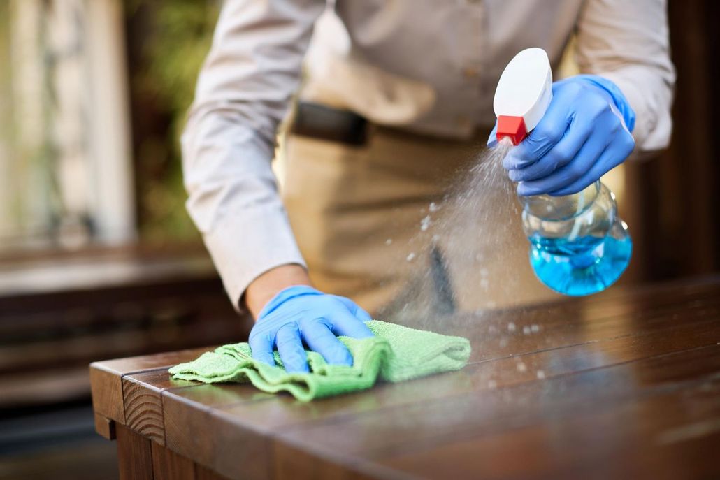 Una persona con i guanti blu spruzza del detergente e pulisce un tavolo di legno con un panno verde.