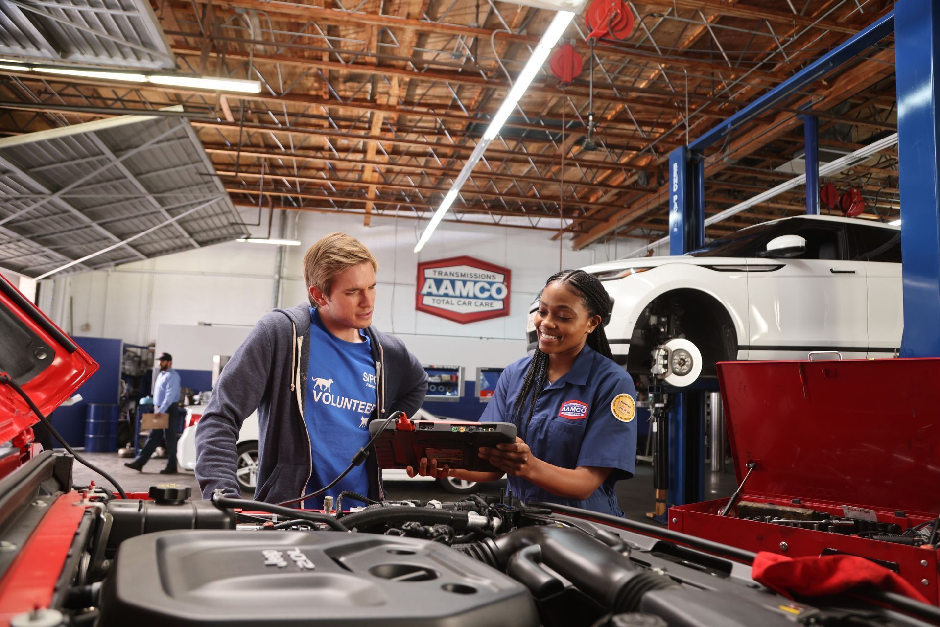 A car mechanic showing a customer a tablet near a red car's open hood in an auto repair shop.