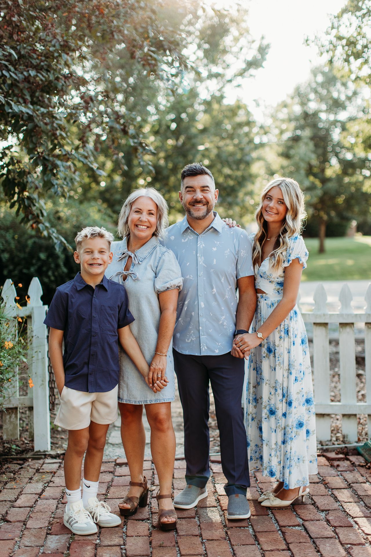 Family of four poses outdoors by a white picket fence, holding hands.