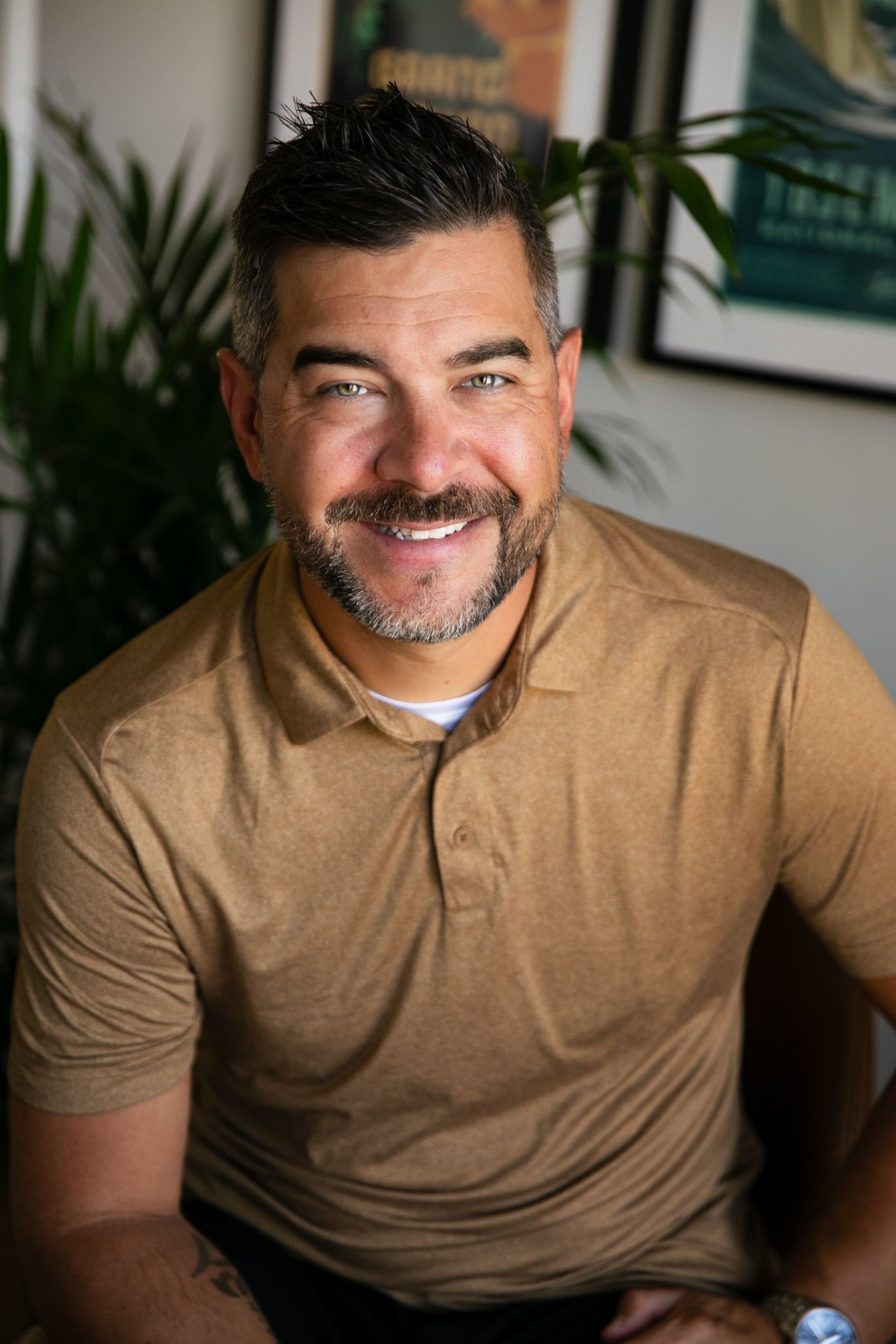 Smiling man with salt and pepper hair, wearing a tan polo shirt, posing indoors with a leafy plant and framed art.