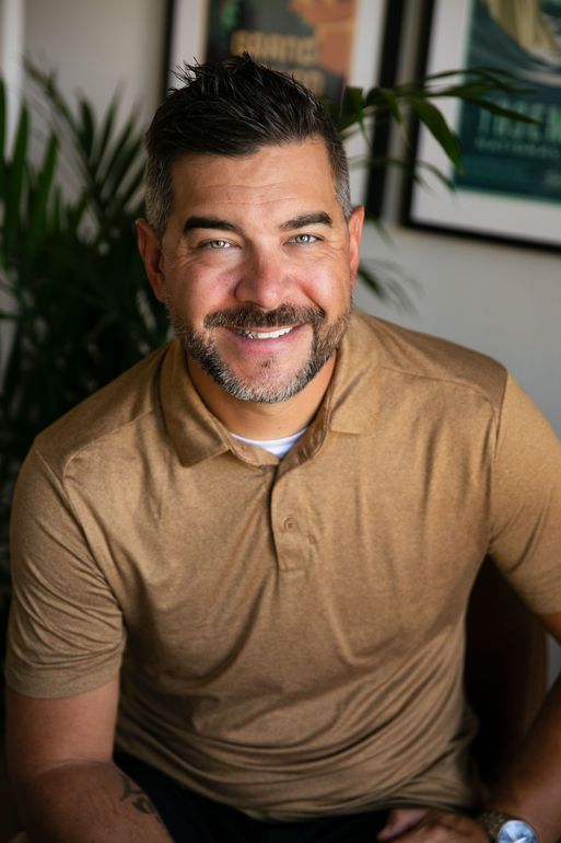 Smiling man with salt and pepper hair, wearing a tan polo shirt, posing indoors with a leafy plant and framed art.