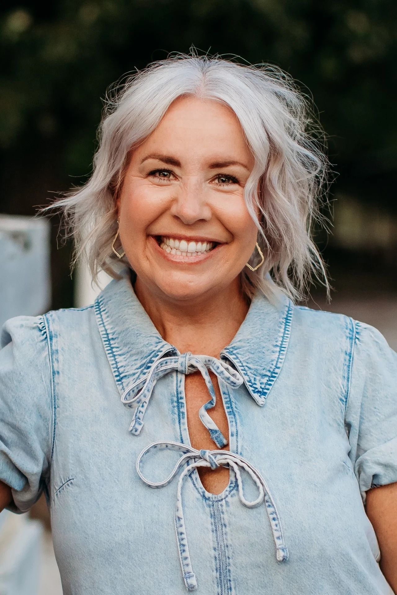 Woman with gray hair smiles, wearing a denim top with a tied neckline; outdoor setting.