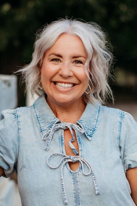 Woman with gray hair smiles, wearing a denim top with a tied neckline; outdoor setting.