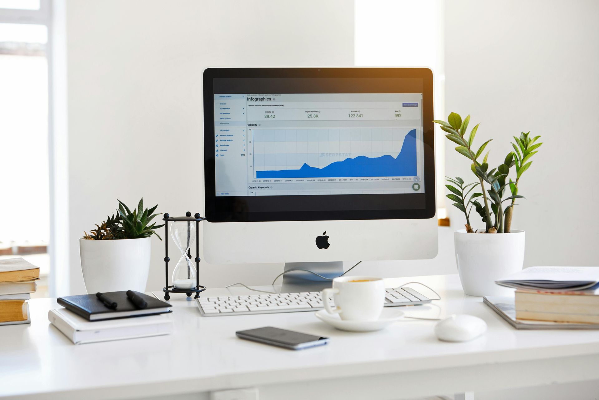 Desk with Apple computer displaying a blue graph, hourglass, plants, coffee, and a phone.