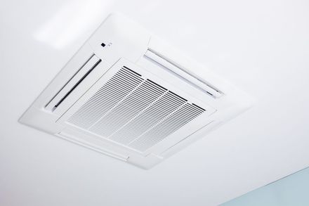White ceiling-mounted air conditioning unit with vents and a grid, set against a white ceiling.