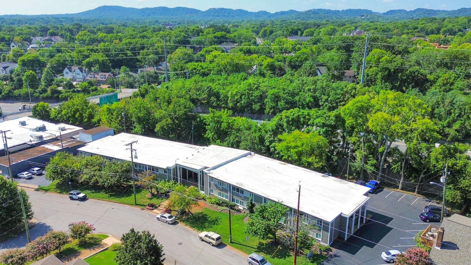 Aerial view of a white-roofed building surrounded by trees and parking, with mountains in the distance