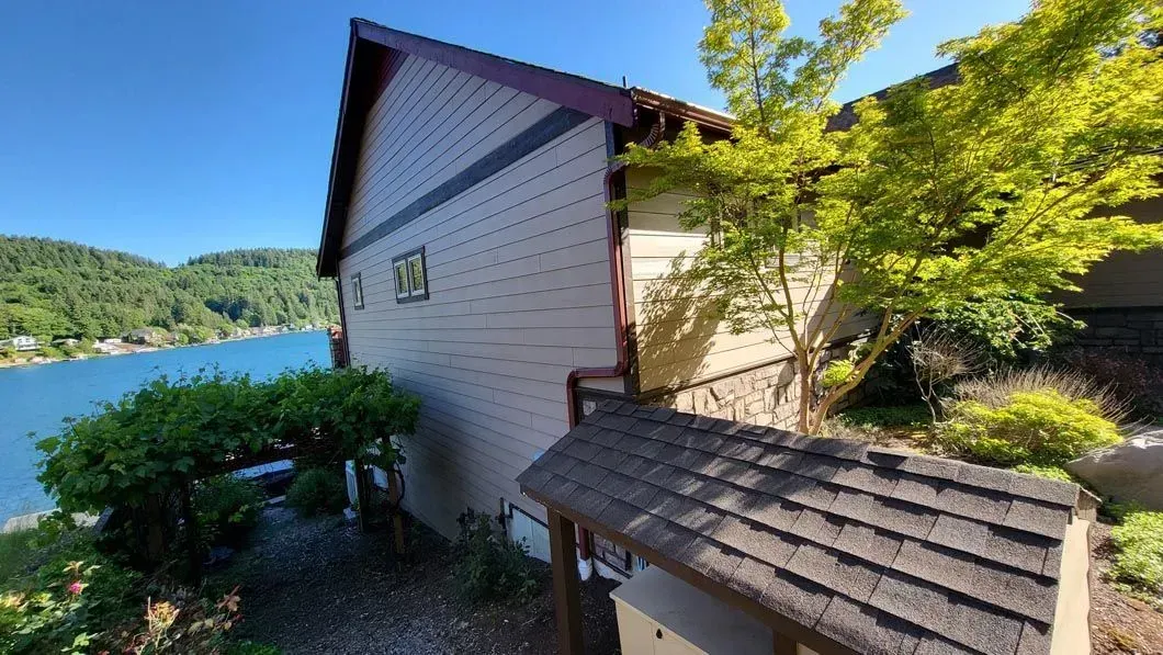 Lakeside cabin with shingled roof, surrounded by trees and bushes beside blue water and a sunny hillside
