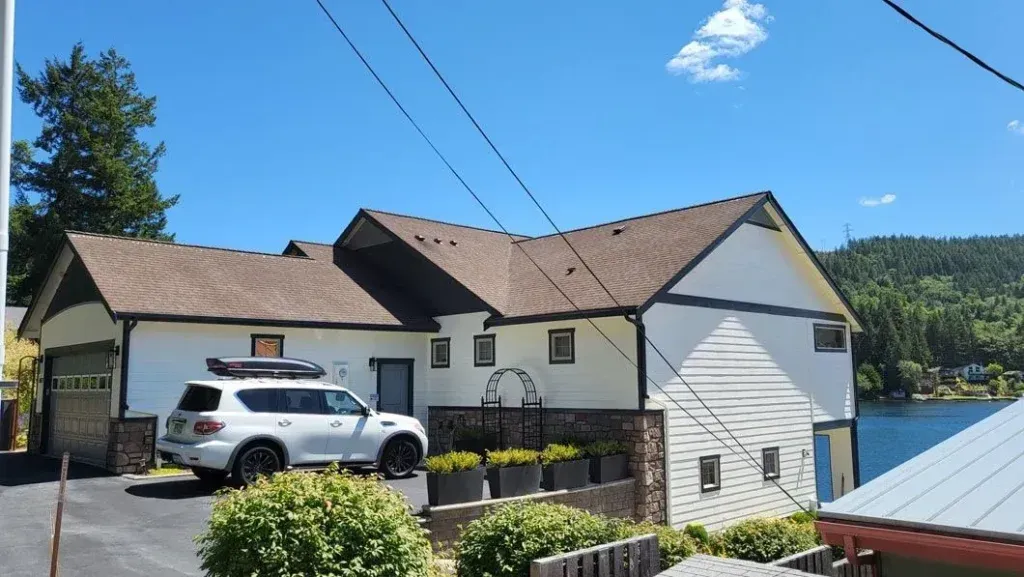 White house with brown roof by a lake, white SUV parked outside, blue sky and trees in the background