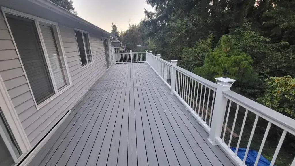 Long gray deck beside a house with white railings and trees beyond