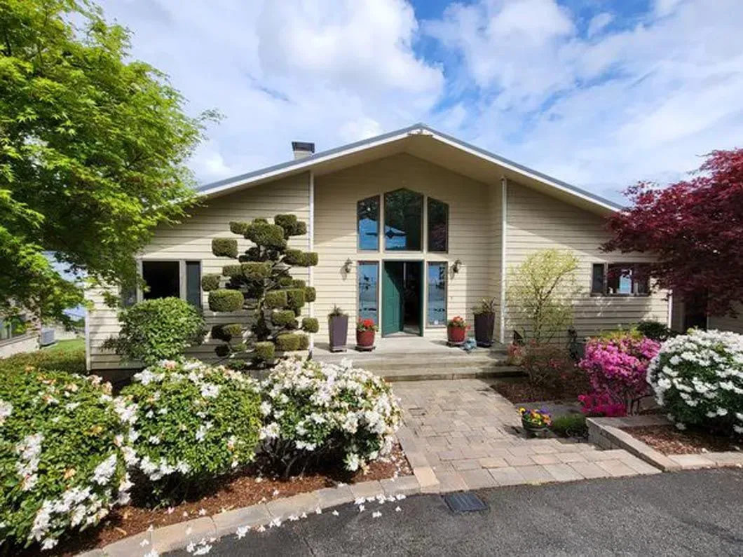 Modern house with large windows, stone walkway, and colorful flowering garden under a blue sky