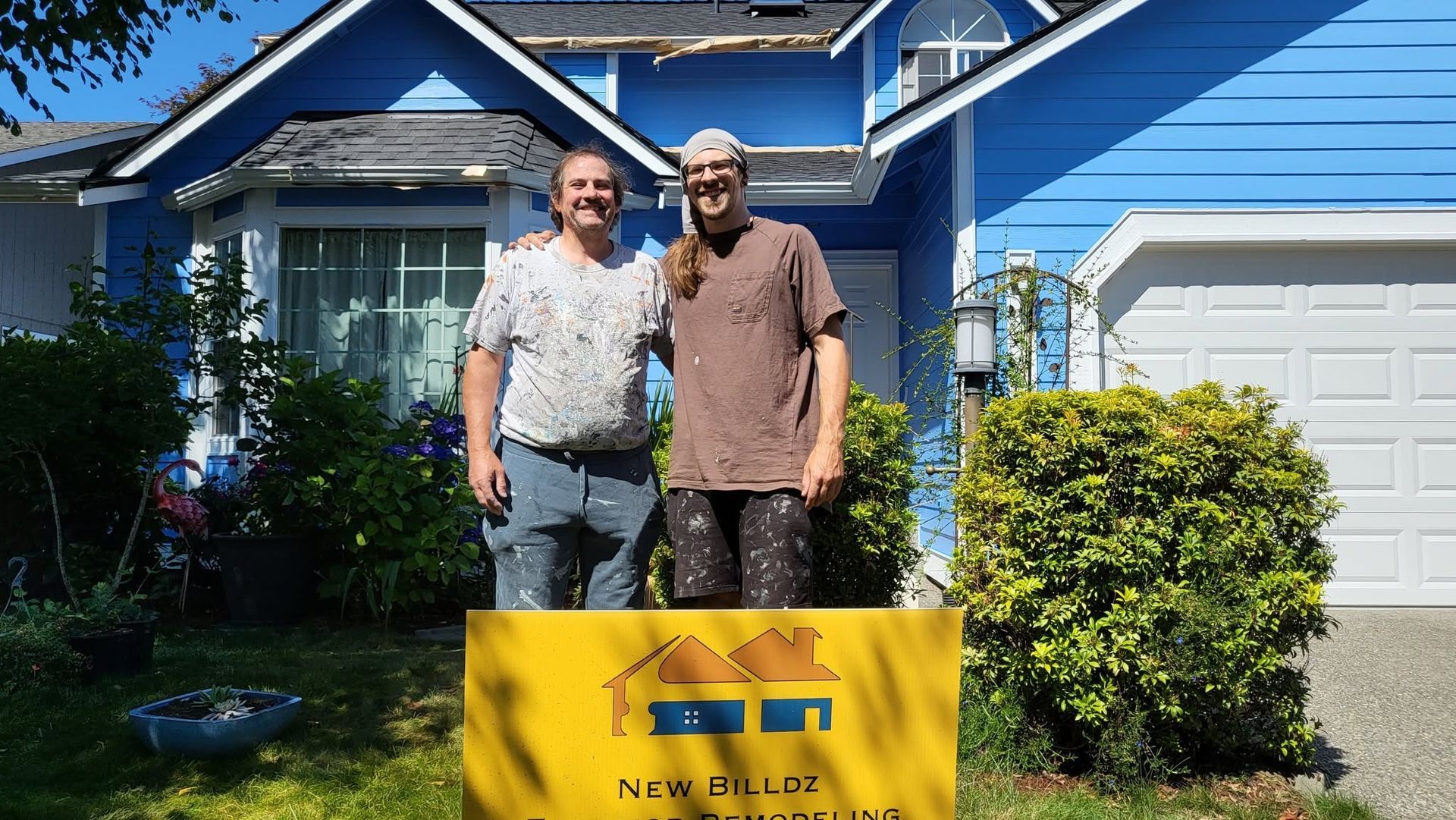Two people standing in front of a blue house, holding a yellow sign in the front yard.