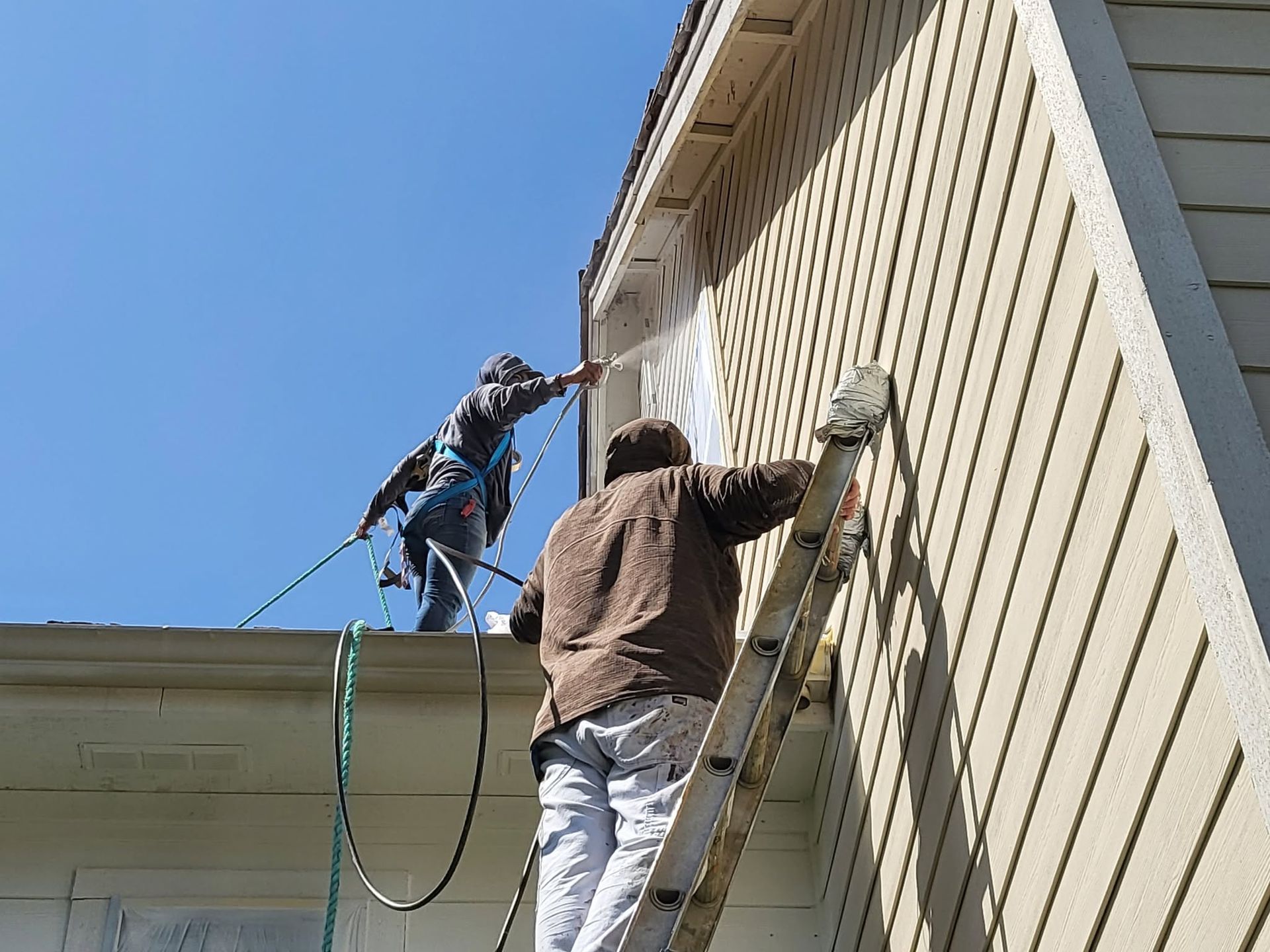 Two workers pressure-wash the side of a house from a ladder and roof.