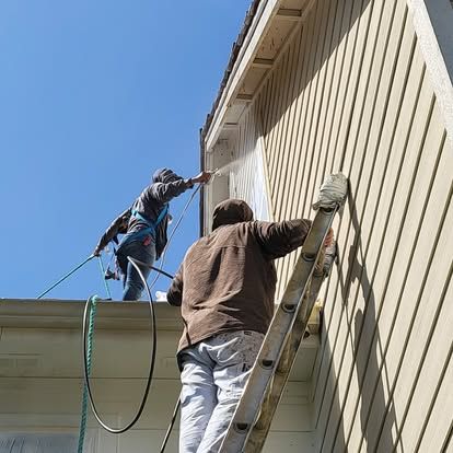 Two workers on a ladder and roof edge painting the side of a beige building under a blue sky