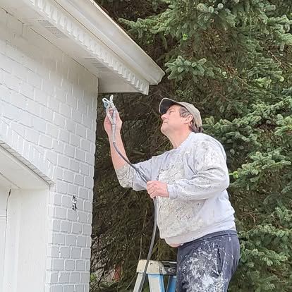 Person painting the exterior trim of a white house beside tall trees