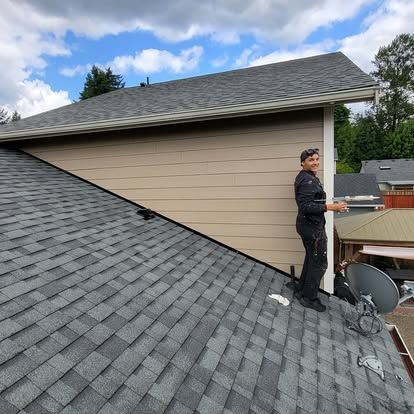Worker on a shingled roof inspecting a house exterior beside a small rooftop addition.