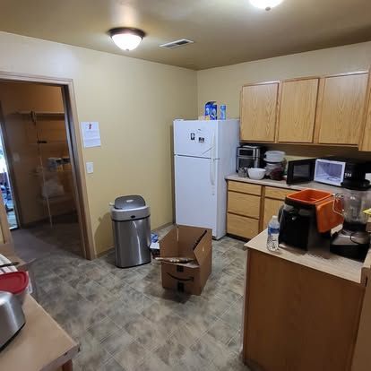 Small kitchen with white refrigerator, wooden cabinets, trash can, and boxes on the floor.