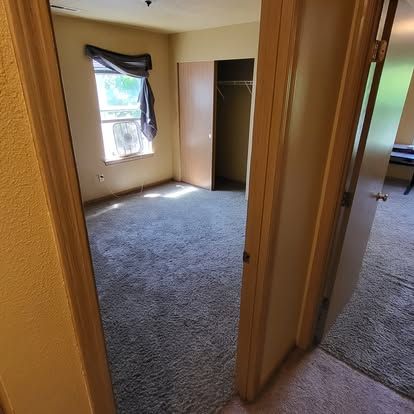 Hallway view into two carpeted bedrooms with beige walls, open doors, and a window with a dark curtain