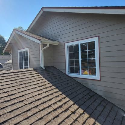 Two-story house exterior with tan siding, white-trimmed windows, and a shingled roof in bright sunlight
