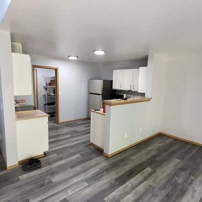 Empty modern kitchen and dining area with gray flooring, white cabinets, and stainless steel refrigerator.