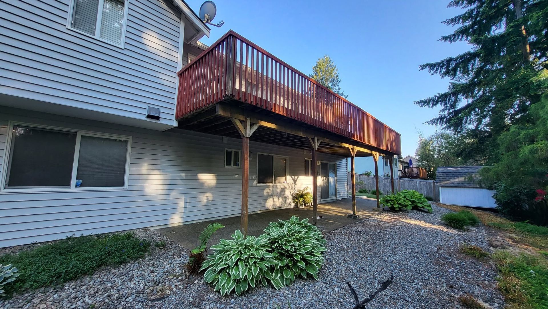 Backyard patio with red wooden deck, gravel ground, house siding, and surrounding greenery