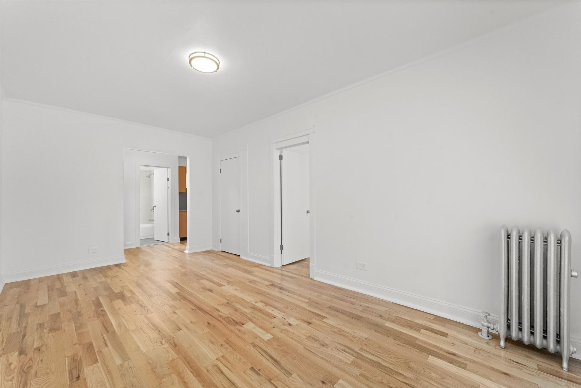Empty room with hardwood floors, white walls, two doors, and a radiator.