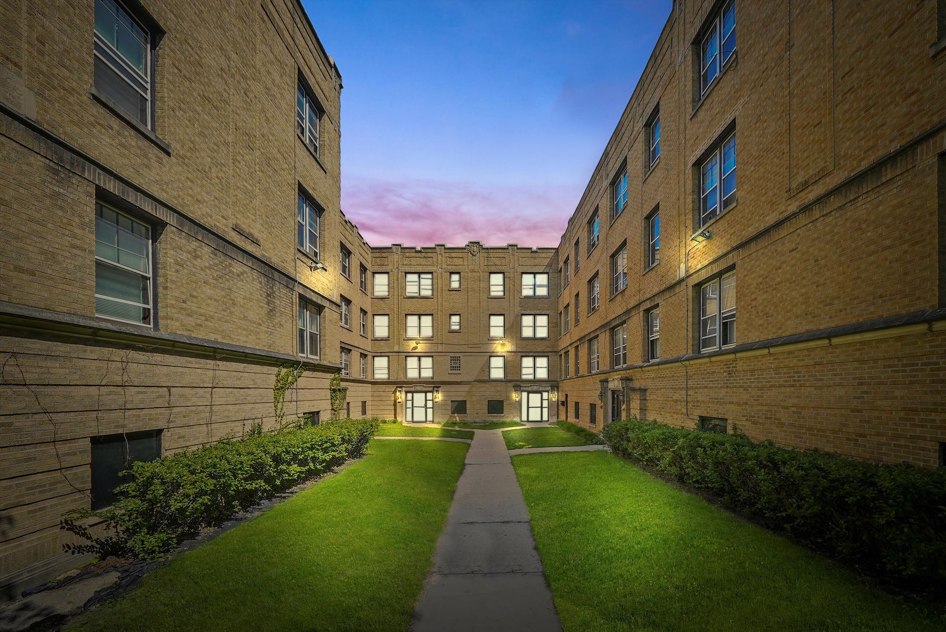 Apartment buildings face each other, framing a courtyard with green grass and a pathway. Dusk sky.