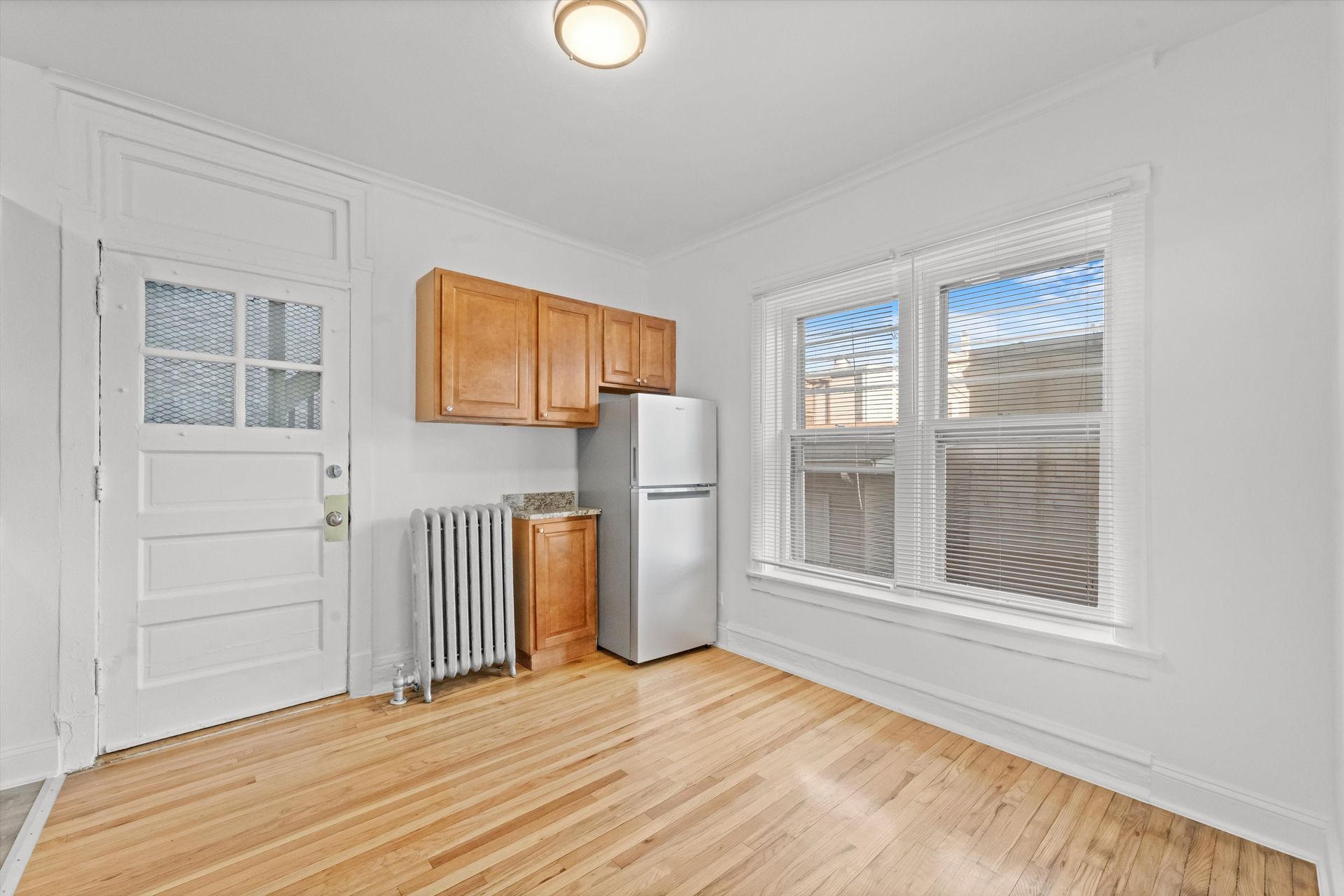 Small kitchen with wooden floor and cabinets, window, and fridge. White walls and door.