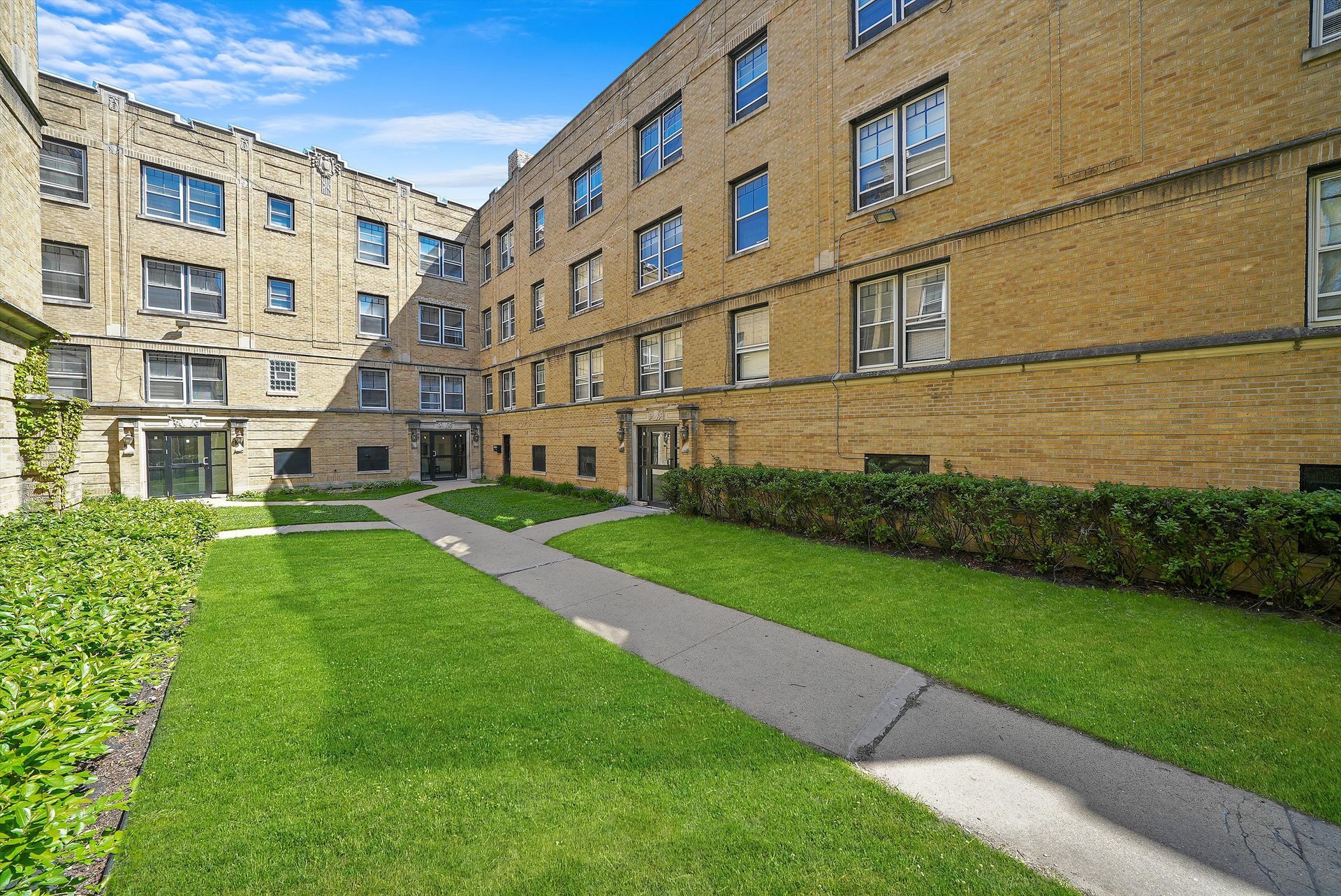 Courtyard of a tan brick apartment building with green grass, a walkway, and bright blue sky.