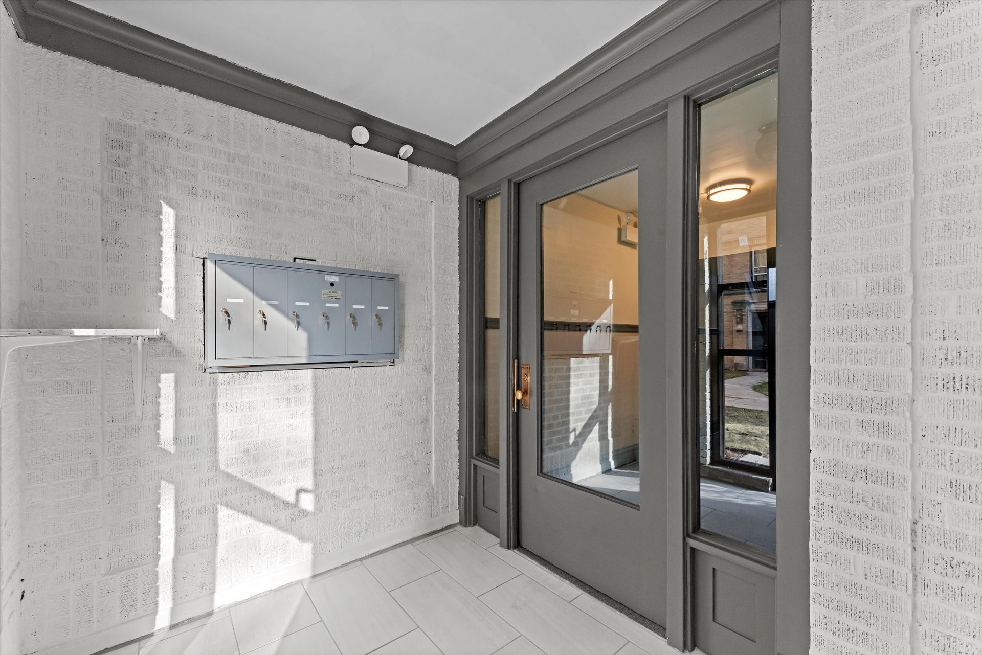 Apartment entryway with white brick walls, gray door and trim, and mailboxes.