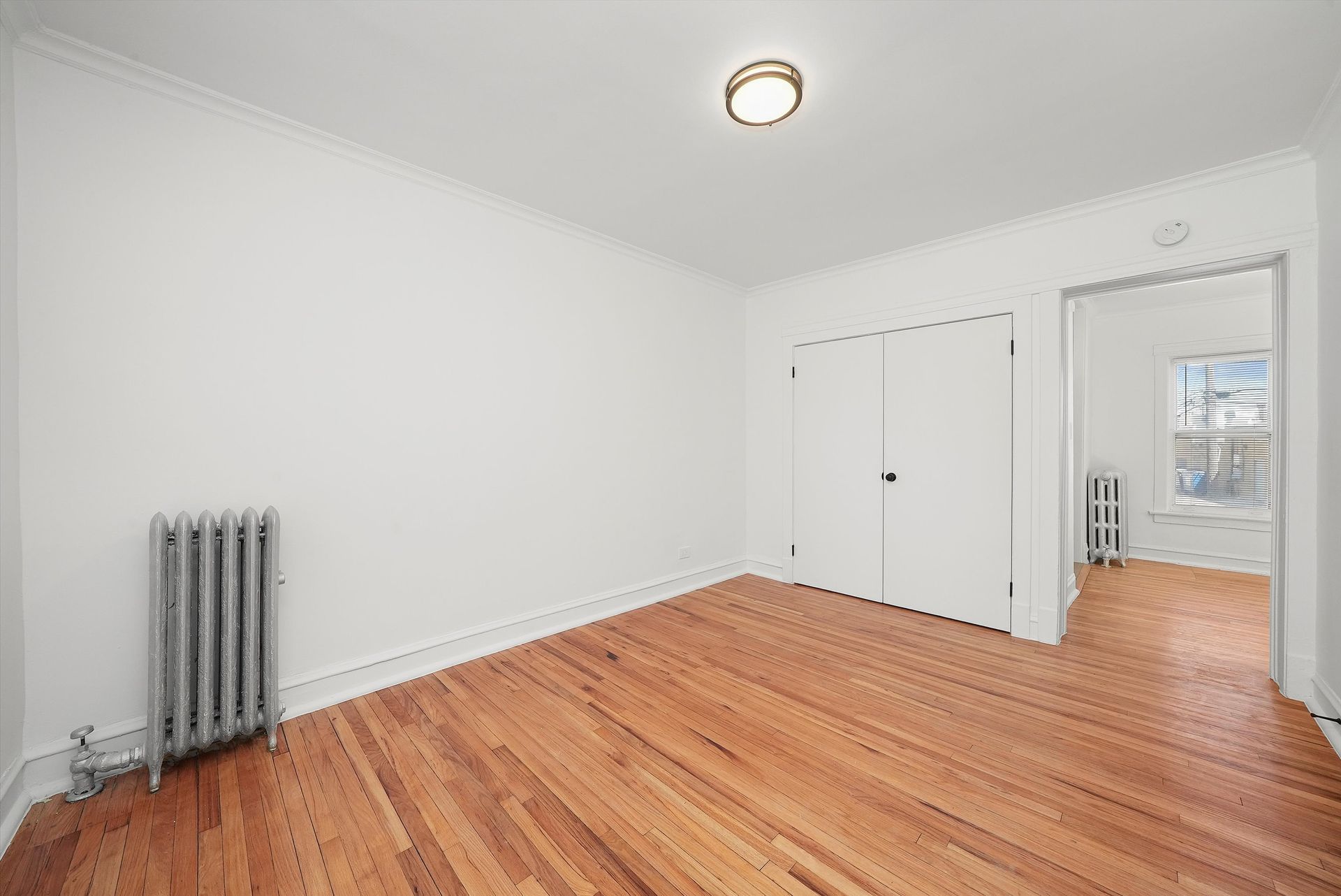 Empty room with hardwood floors, white walls, closet, and radiator. View into another room with a window.