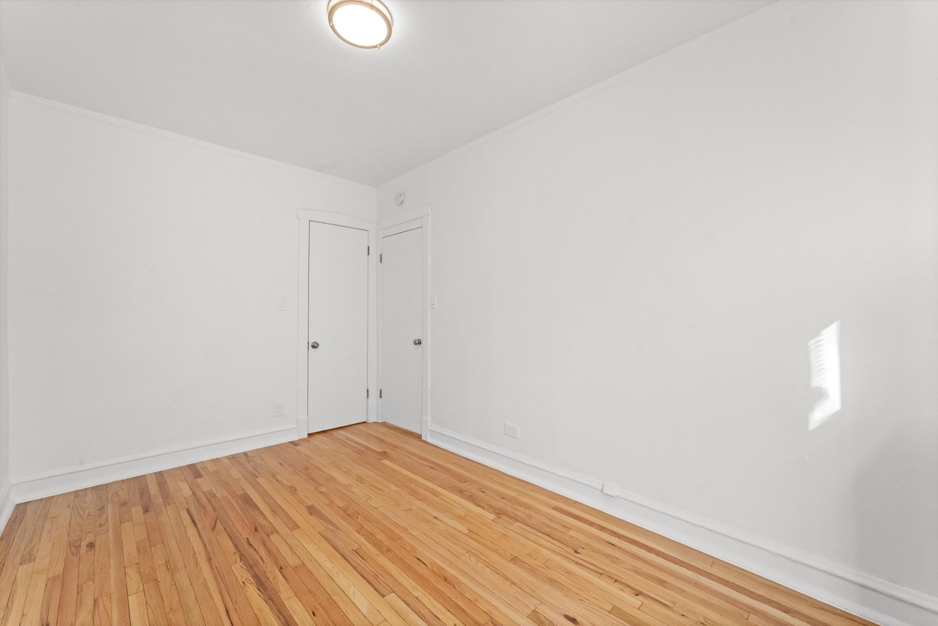 Empty room with hardwood floors, white walls, two doors, and a ceiling light.