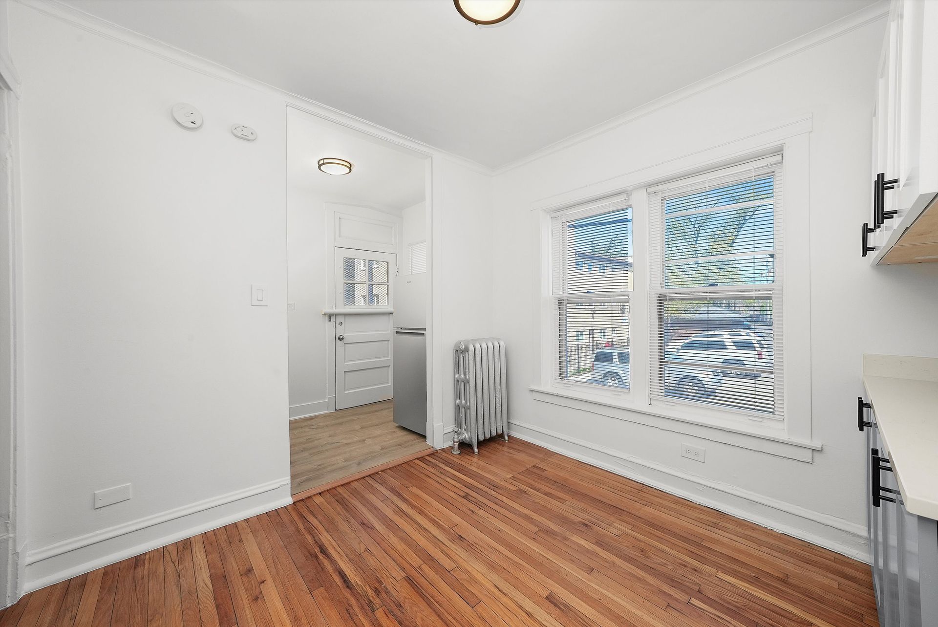 Empty white room with hardwood floor, window, and doorway to kitchen.