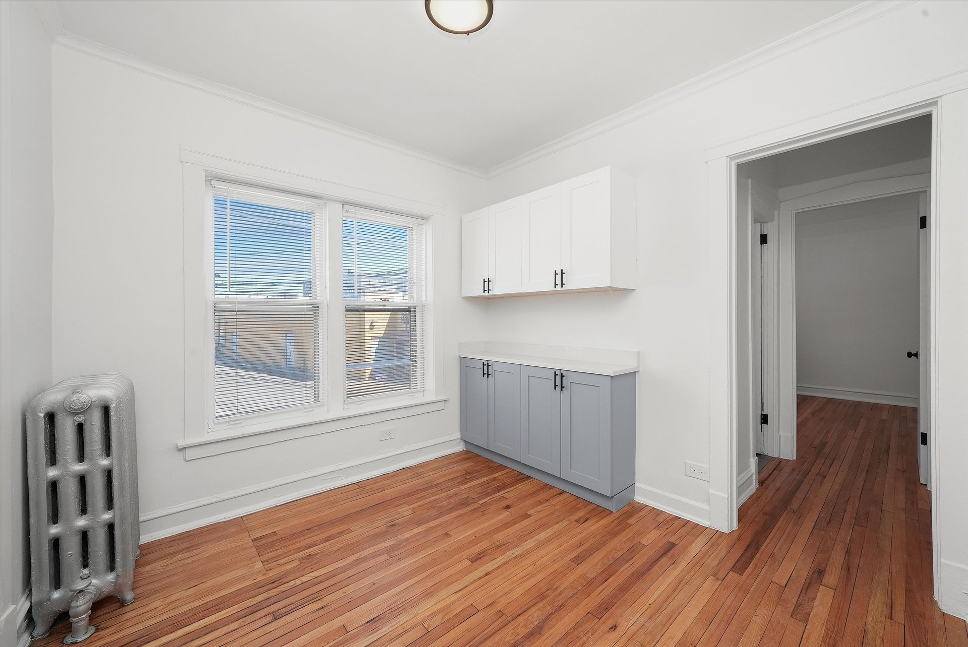 Empty room with hardwood floors, window, radiator, and cabinets.