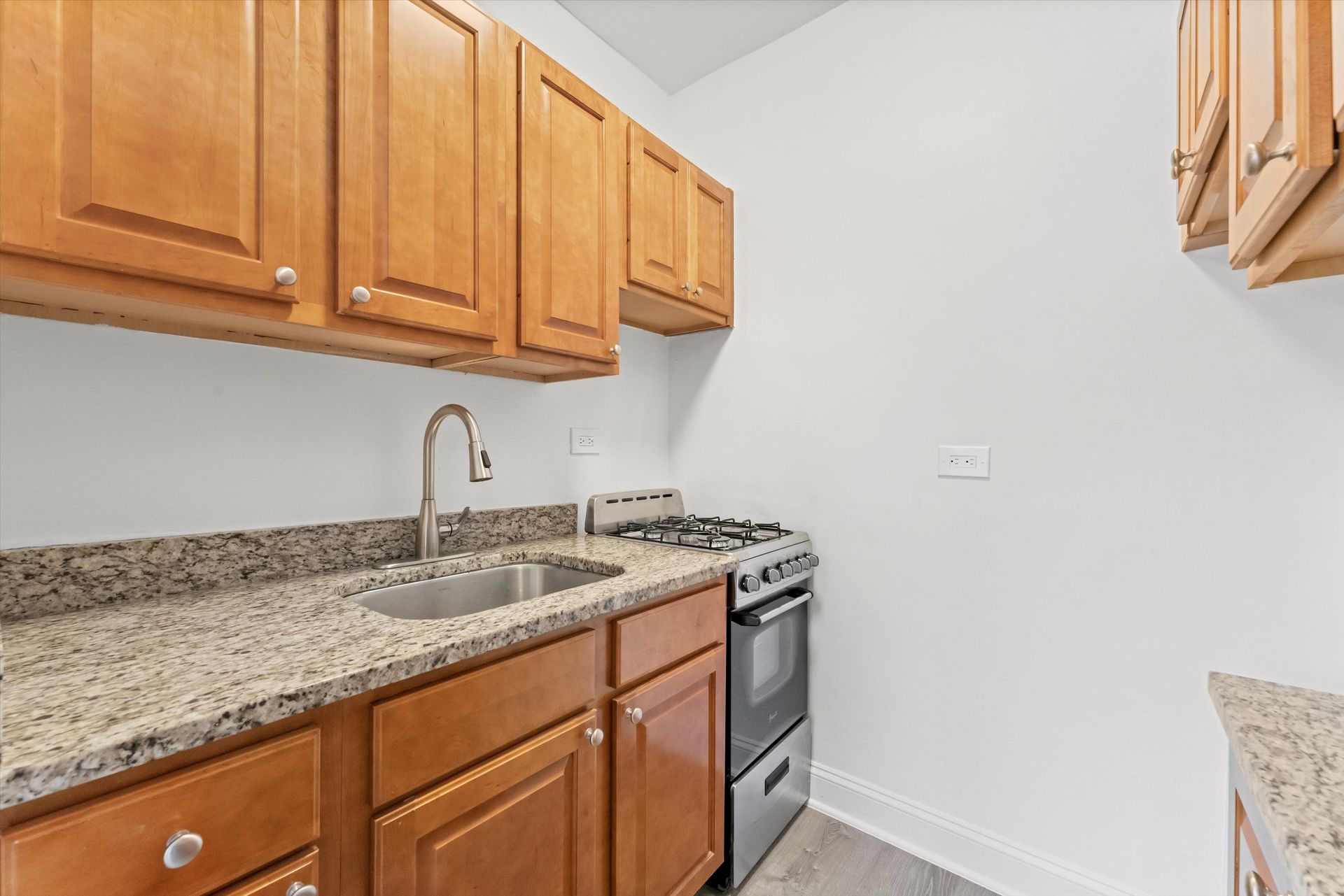 A long, narrow kitchen with white cabinets, appliances, and beige countertops and floor. Doorway at end.