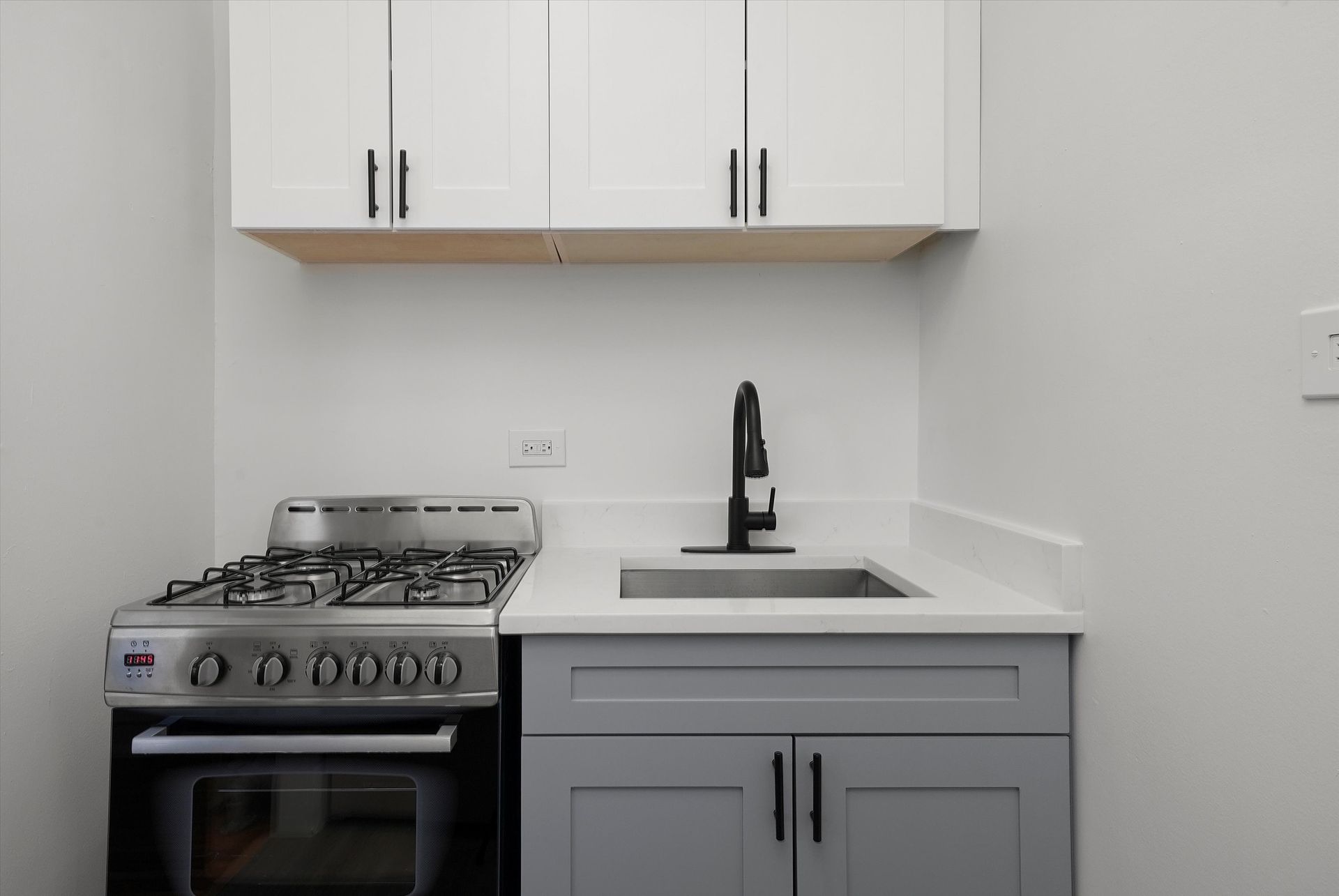 Small kitchen with a stove, sink, and cabinets. White walls, gray and white cabinets, black faucet.