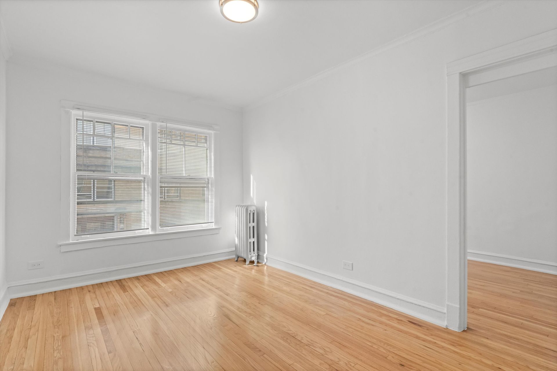 Empty room with hardwood floors, white walls, window with blinds, and doorway.