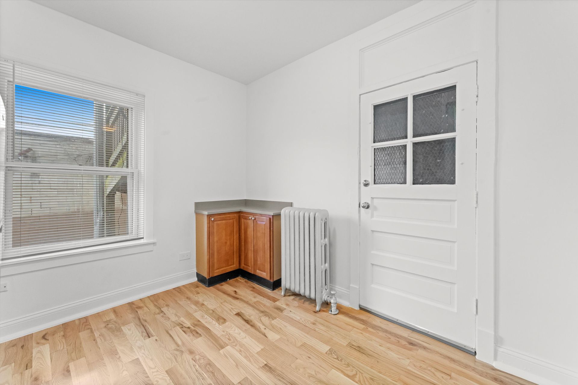 Empty room with hardwood floors, a window, and a door. Includes a small cabinet and radiator.