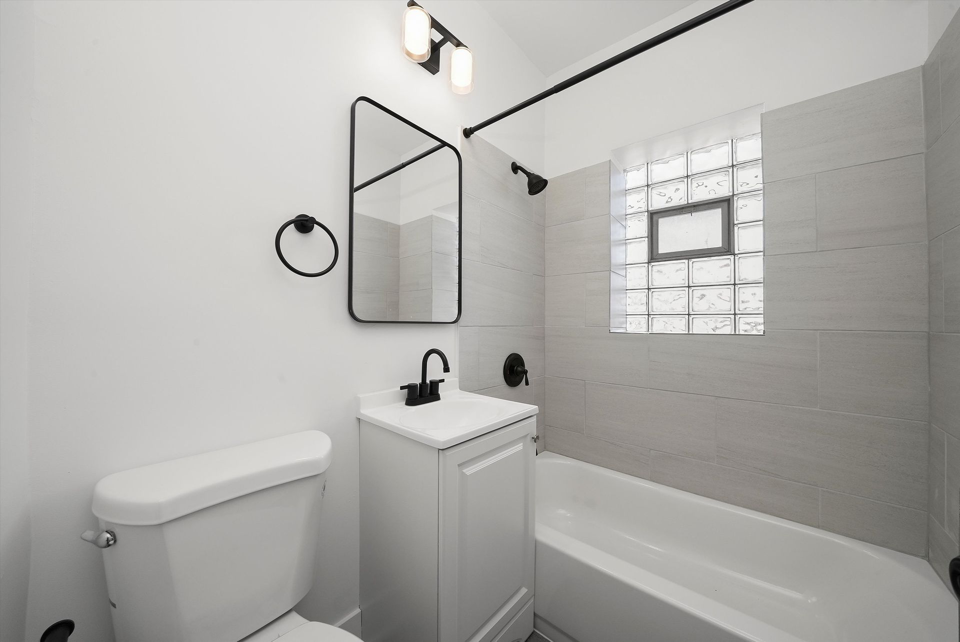 White bathroom with toilet, vanity, mirror, tub, and window. Black fixtures and light.