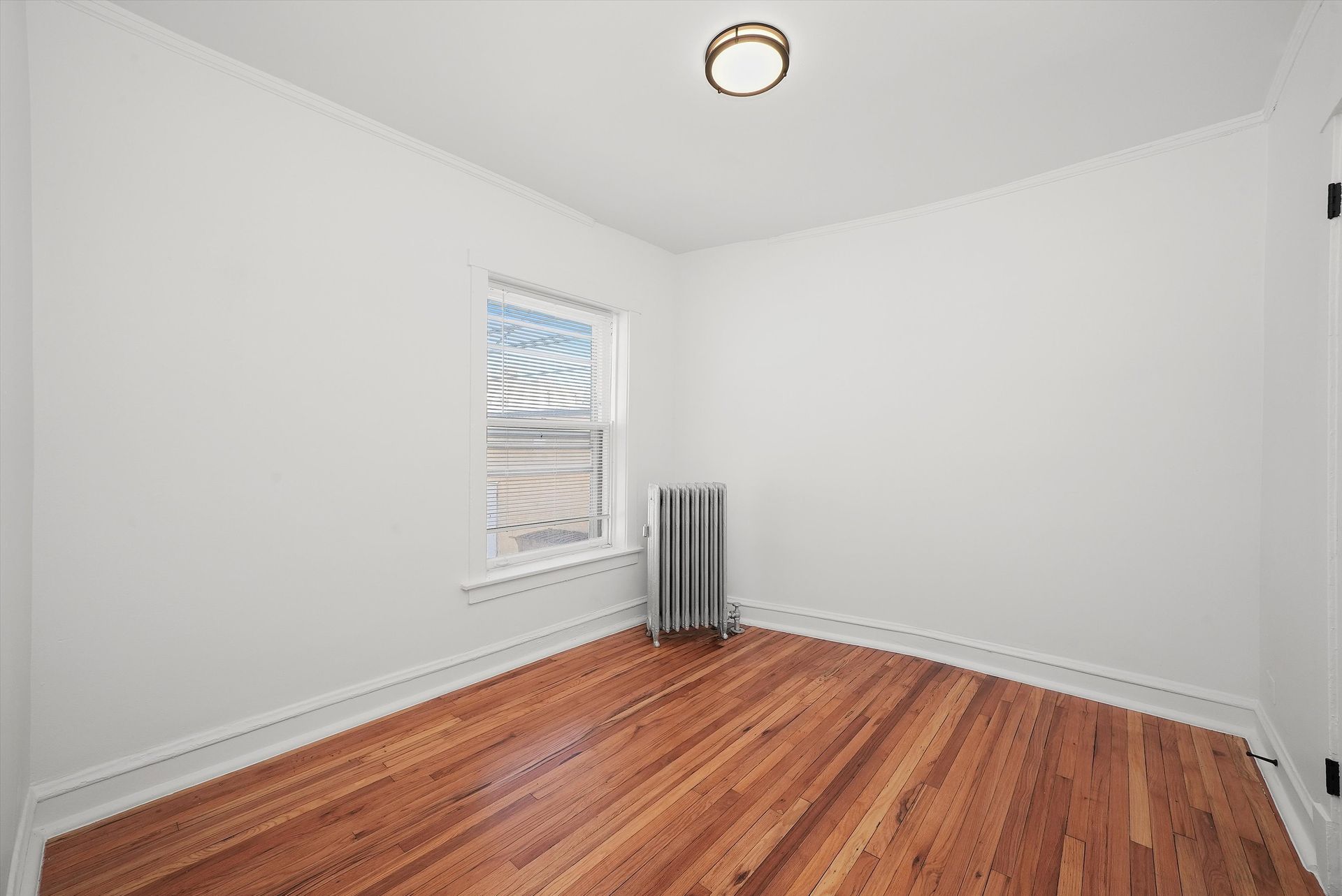 Empty room with hardwood floors, small window, radiator, white walls, and a ceiling light.