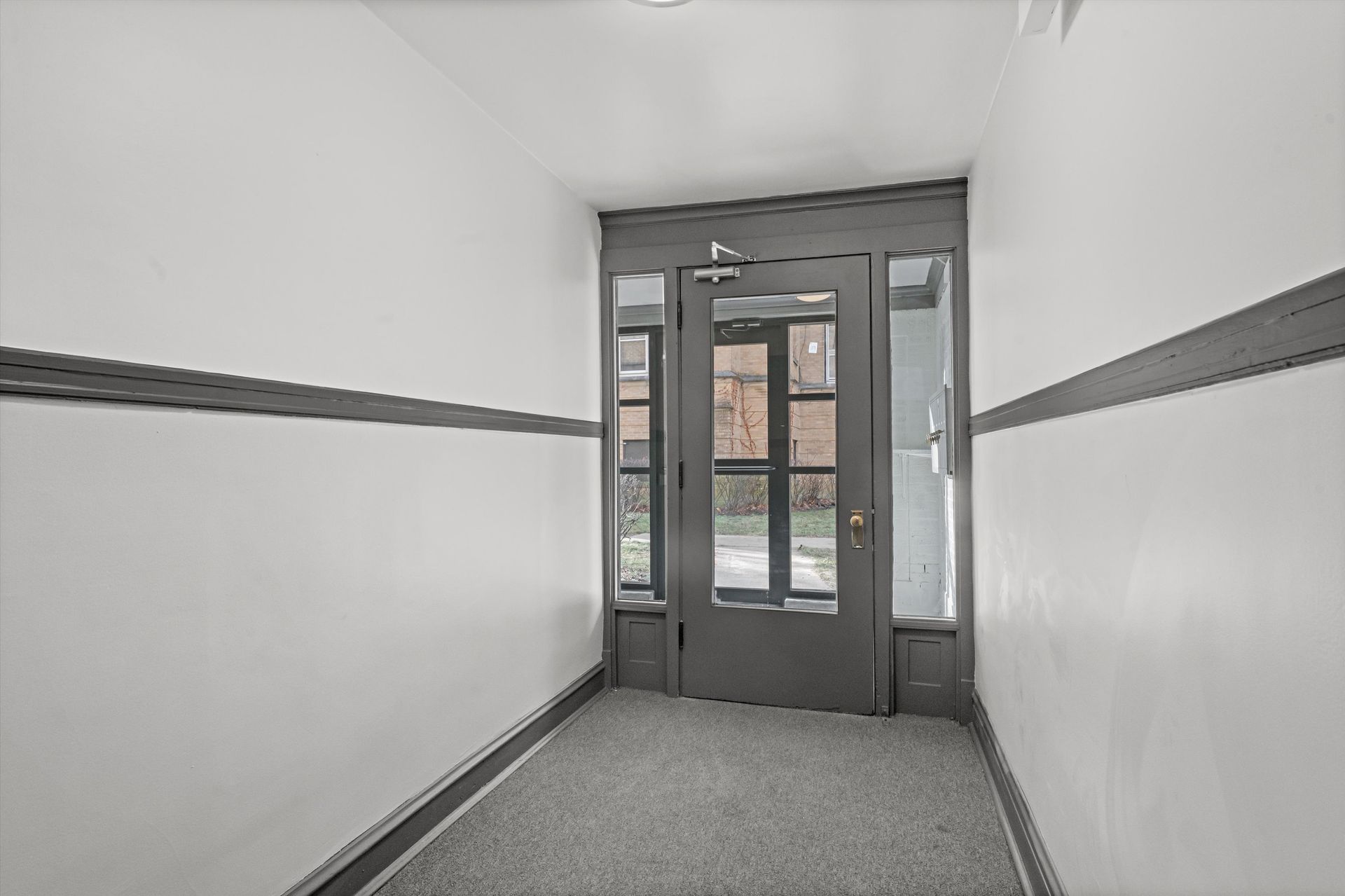 Narrow hallway leading to a gray door with glass panels. White walls with gray trim and carpet.