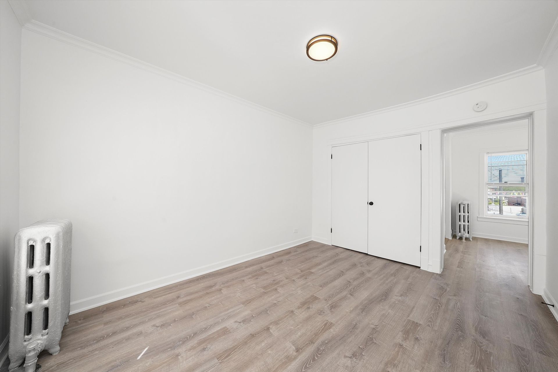 Empty room with white walls, wood floor, radiator, two closet doors, doorway to another room.