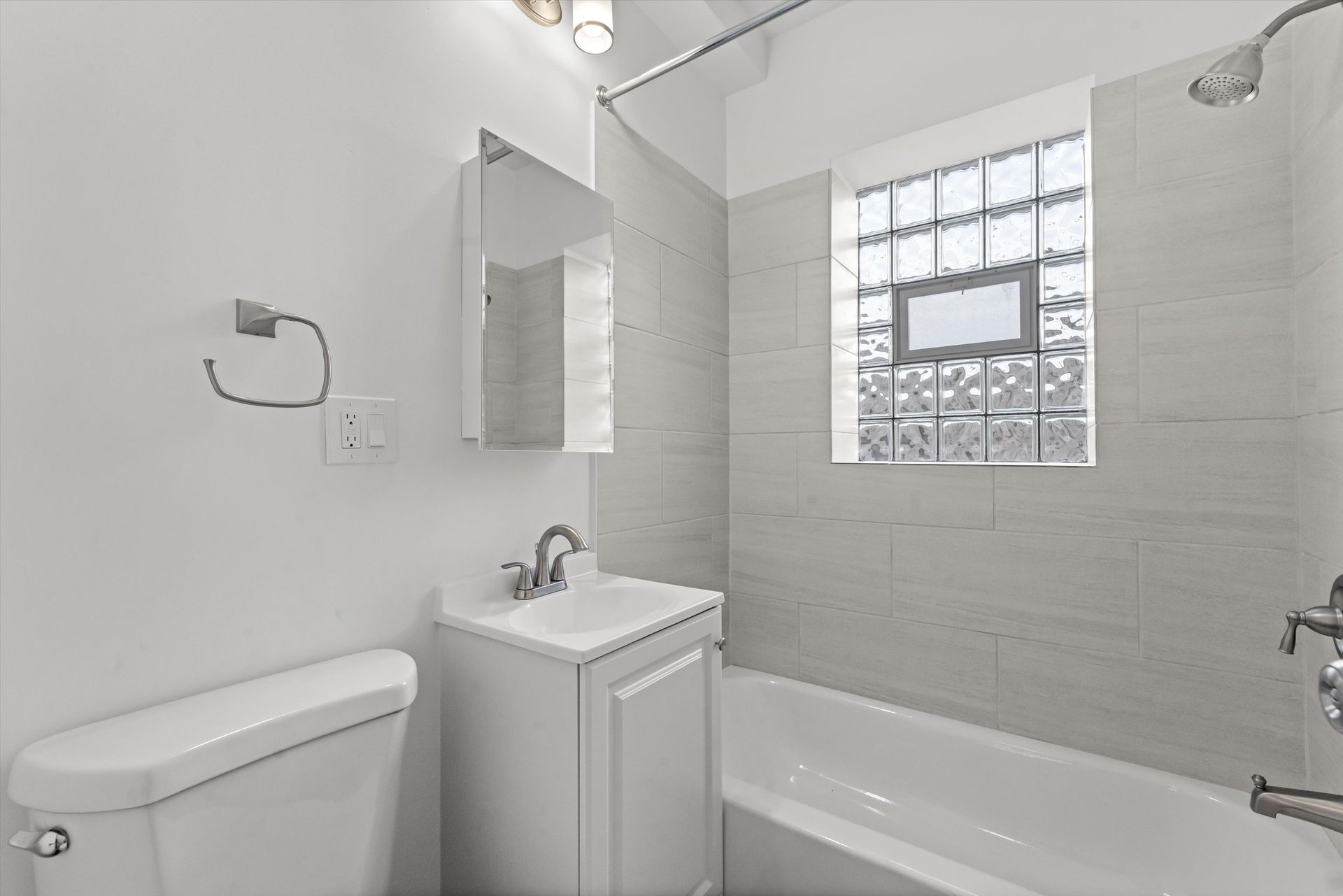 A white bathroom with a toilet, sink, bathtub, and showerhead, next to a window with square glass blocks.