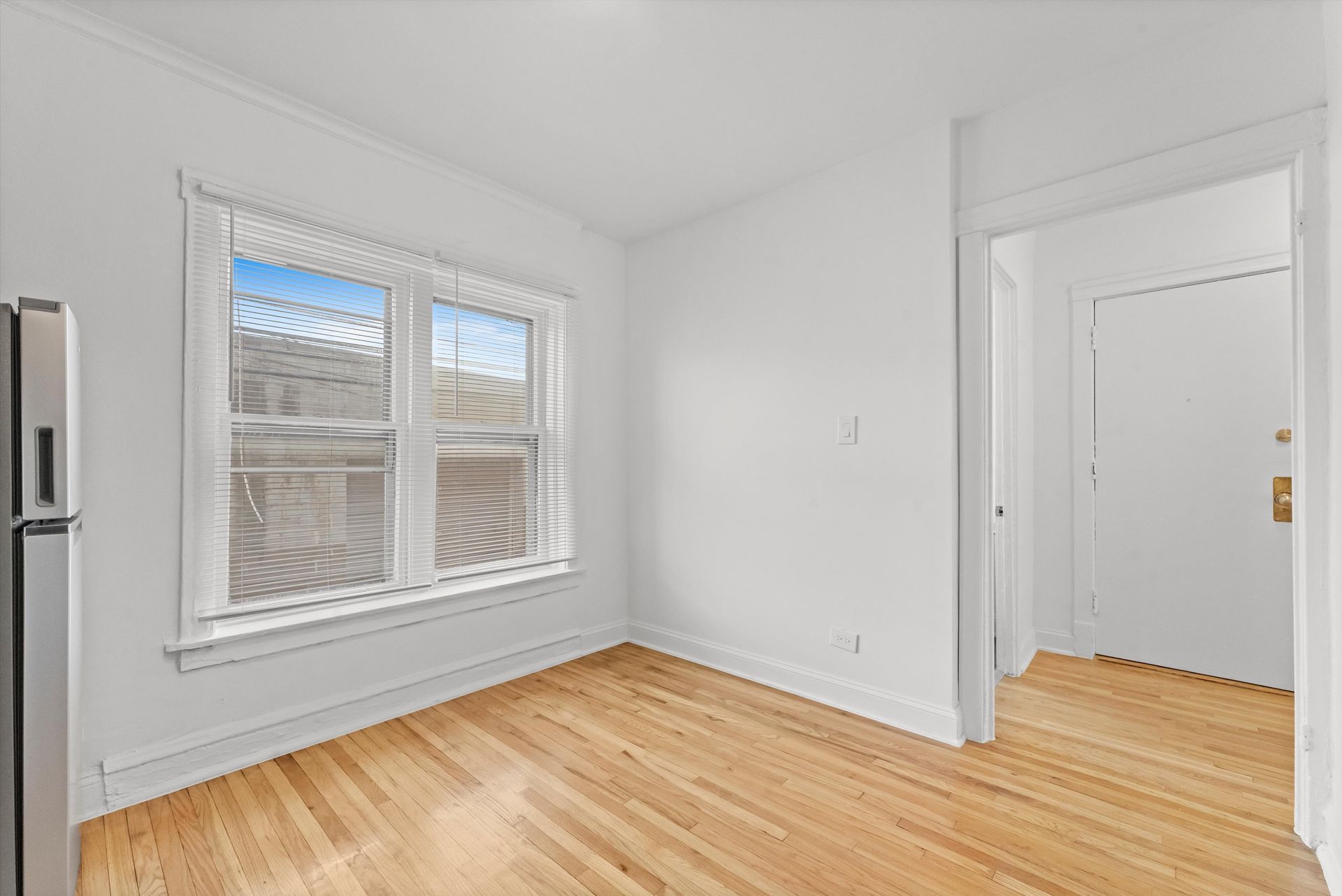 Empty room with hardwood floors, window with blinds, and doorway leading to a white door.