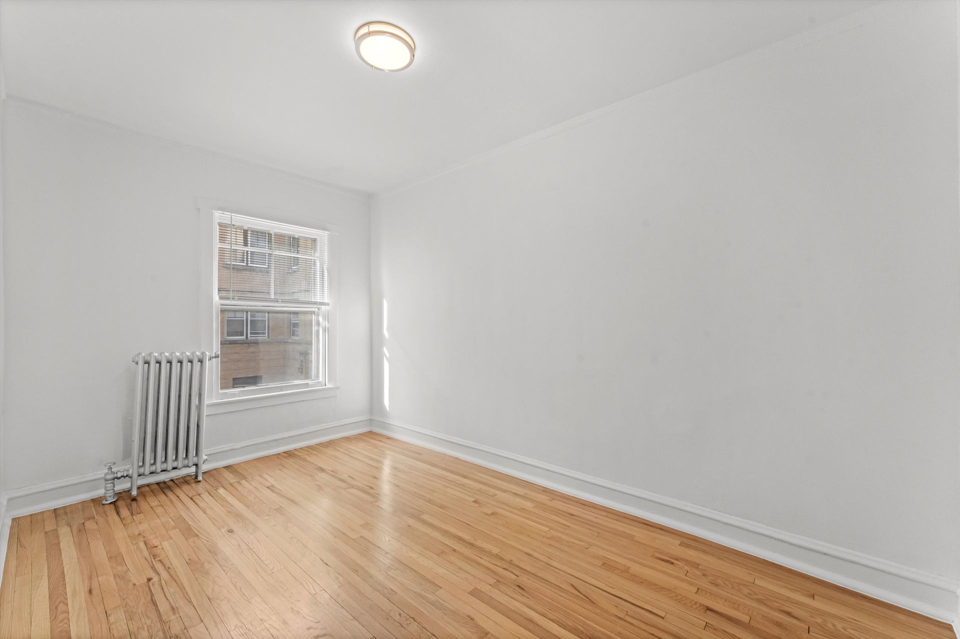 Empty room with wood floor, window, radiator, and white walls.