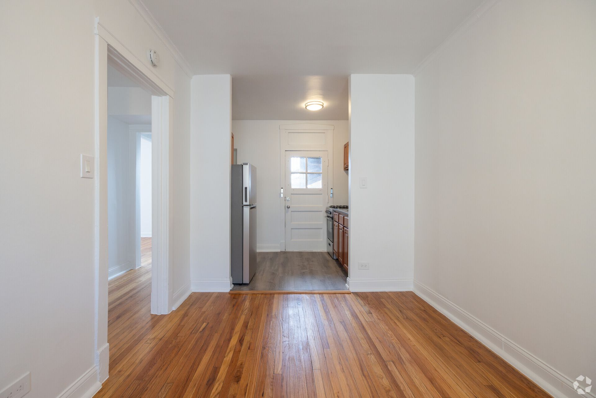 Interior view of apartment, showing hardwood floors, white walls, and kitchen entrance.
