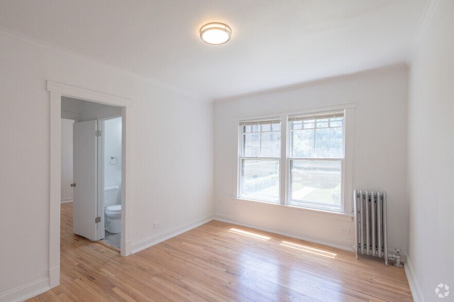 Empty room with hardwood floors, white walls, and a window; doorway to a bathroom.