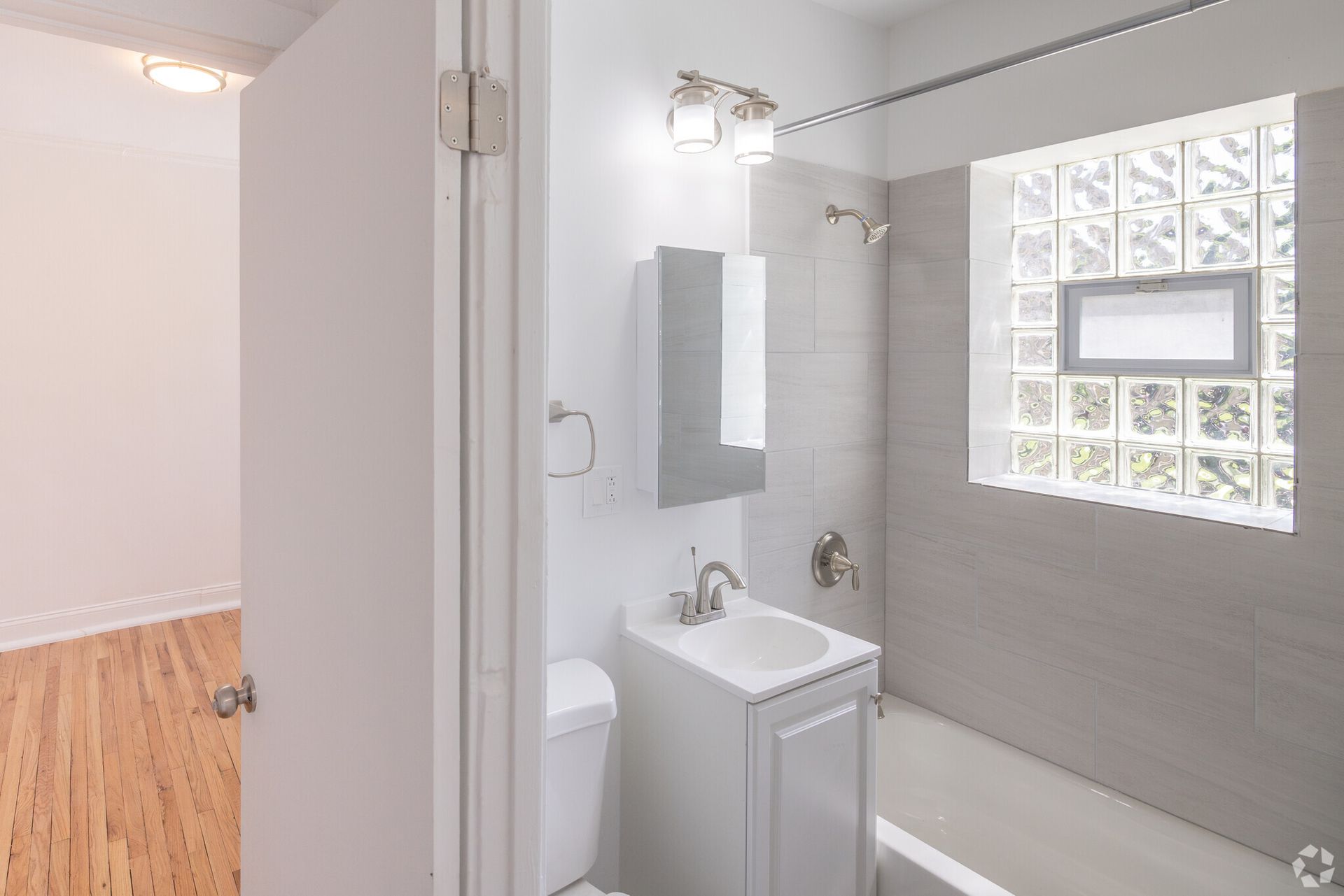 Bathroom with white fixtures and gray tile. View into a hallway with hardwood floors.