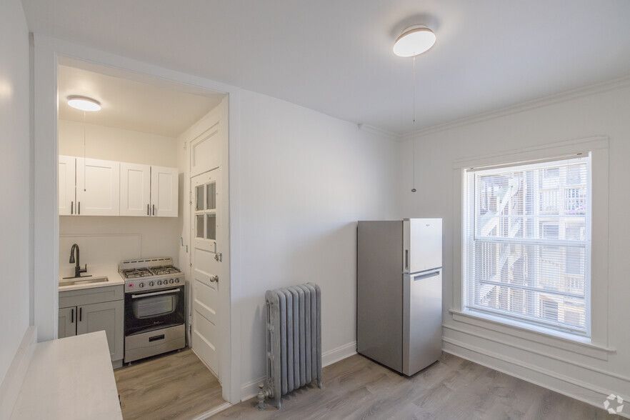 Small apartment kitchen with refrigerator, stove, cabinets, window, and radiator. White walls, gray floors.